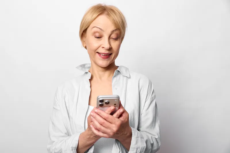 Middle aged woman holding smartphone indoors on plain white background calm smile looking down at screen studio portrait technology mobile communication moment
