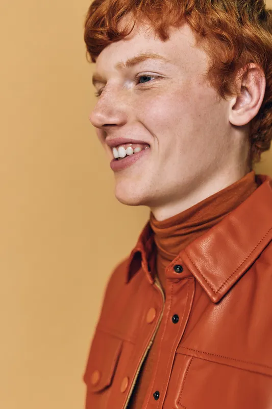 Young man with red hair and freckles wearing orange leather jacket and turtleneck smiling sideways on warm beige background portrait closeup in studio lighting.