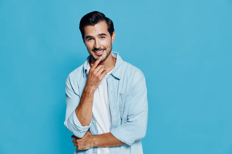 Charming smiling young man with stylish hair in casual light blue shirt, stands thoughtfully against solid turquoise background. Engaging and lighthearted concept.