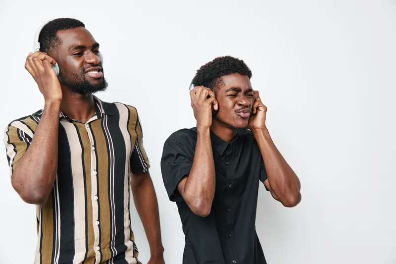 Two African American men with dark skin listening to music on headphones, enjoying sound and rhythm with closed eyes and relaxed expressions on white background.