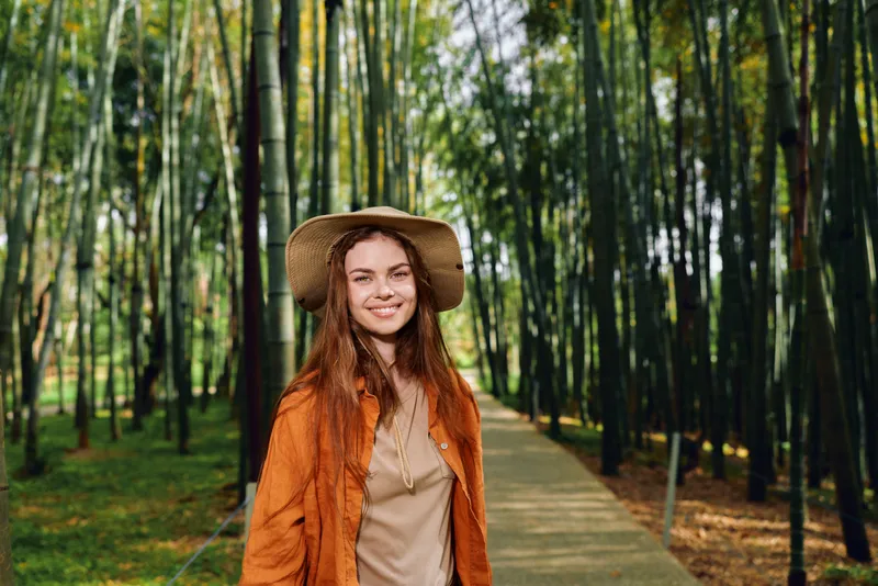 Woman in a straw hat with a warm smile in a bamboo forest on a wooden path, portrait of a happy traveler wearing orange jacket, nature walkway with sunlight through tall green stalks.