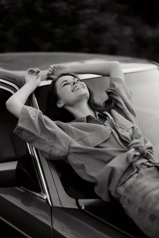 Young woman joyfully relaxing against a vintage car, wearing a casual outfit, with a happy expression, set against a soft, natural background.