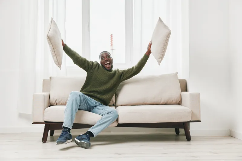 Happy African American man celebrating at home with pillows, wearing casual green sweater and jeans, in a bright, minimalistic indoor setting