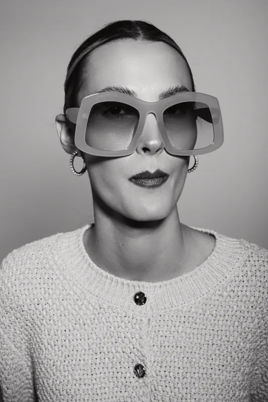 Elegant caucasian woman with pale skin wearing oversized sunglasses and earrings poses in knitted white sweater on grey background.