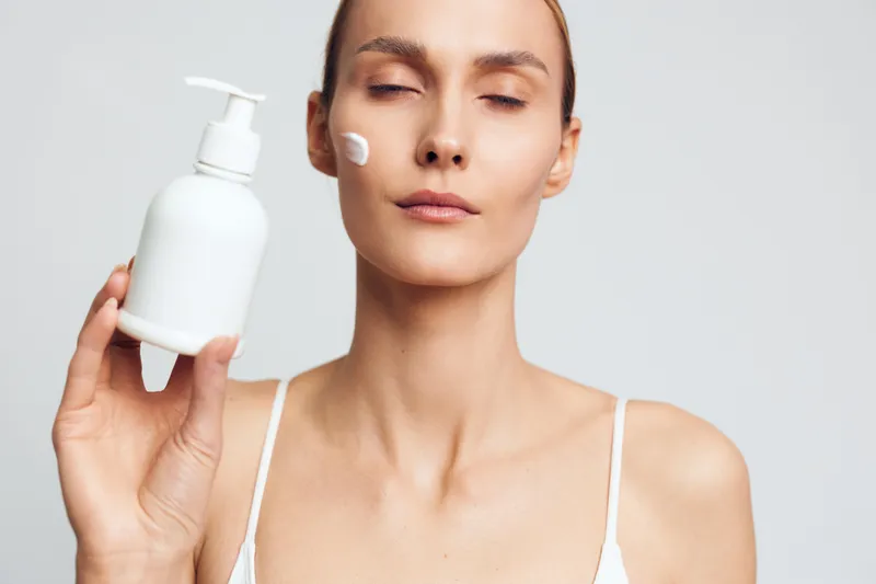 Young woman with clean skin applies white cream on face holds bottle with pump dispenser eyes closed isolated on light background skincare beauty concept.