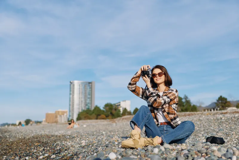 Woman photographer on a pebble beach, wearing a plaid shirt and jeans, sits with camera raised to her eye, sunglasses on, bright day, distant buildings blur on the horizon.