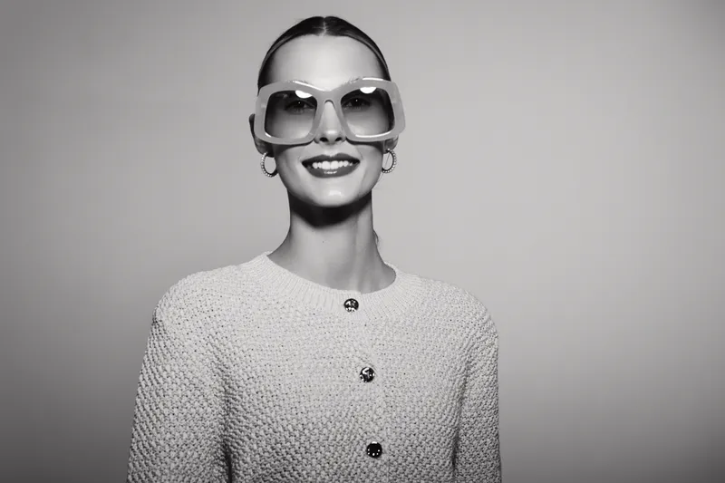 Smiling white woman with fair skin wearing large sunglasses and a textured jacket stands against plain background. Concept of fashion, style, elegance, and confidence.