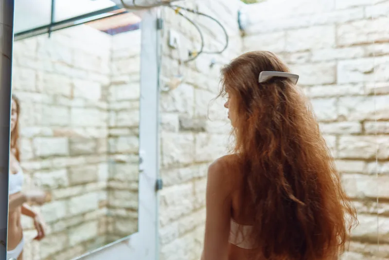 Woman with long hair standing in a rustic shower room with exposed brick walls, natural light, and vintage shower fixtures showing relaxed morning routine.