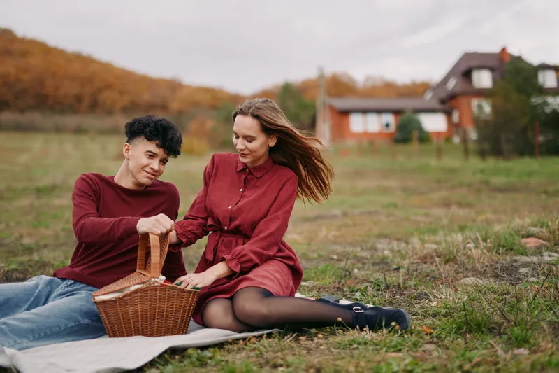 A genuine outdoor picnic scene of a young couple sharing a wicker basket on a blanket in a grassy field, capturing natural interaction, relaxed posture and authentic connection.