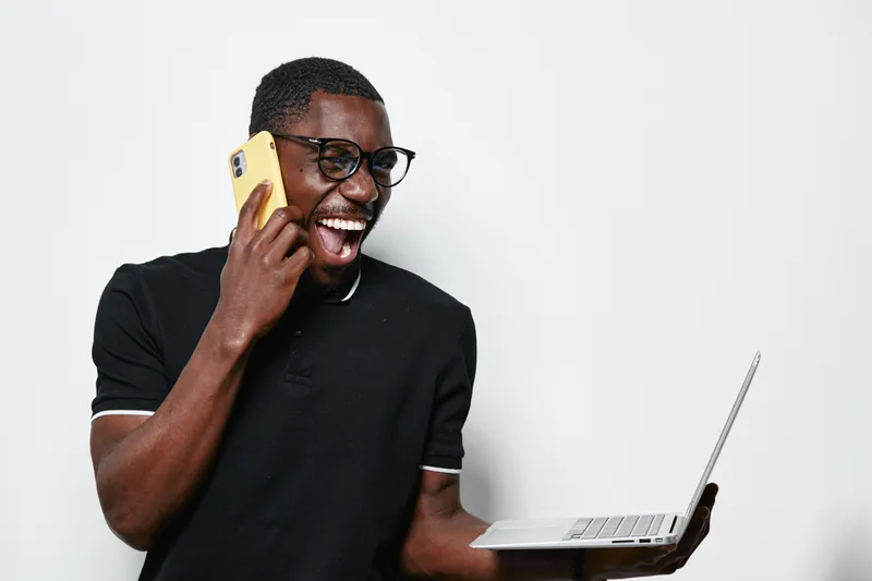 African American man with dark skin talking on phone and holding laptop, smiling and wearing glasses in casual black shirt on white background for communication and technology.