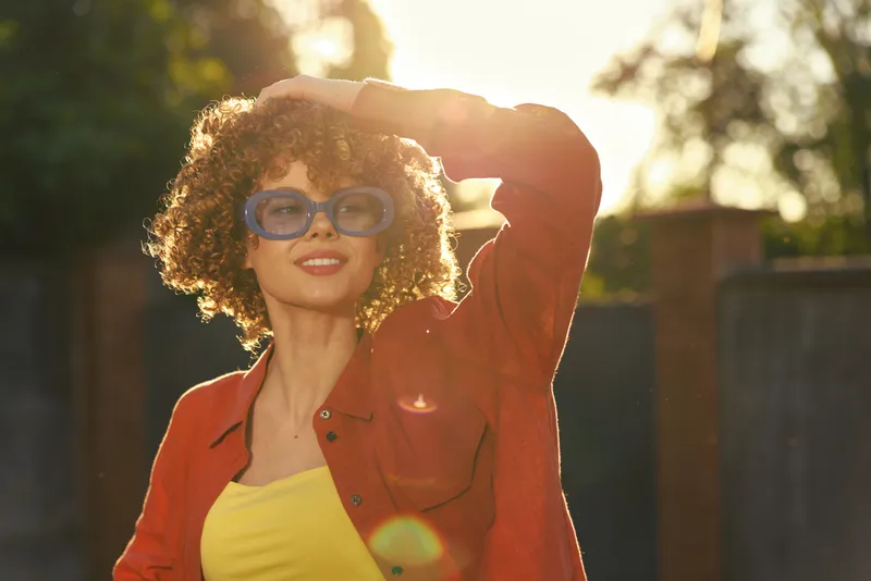 A stylish woman with curly hair wears an orange jacket and yellow top, shielding her eyes from the sun in a sunny outdoor yard. Concept: casual confidence and outdoor fashion