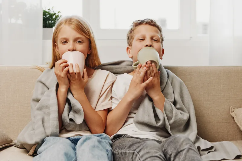 Two happy children wrapped in blankets drink warm beverages sitting on a couch by the window. Cozy home atmosphere with natural light.