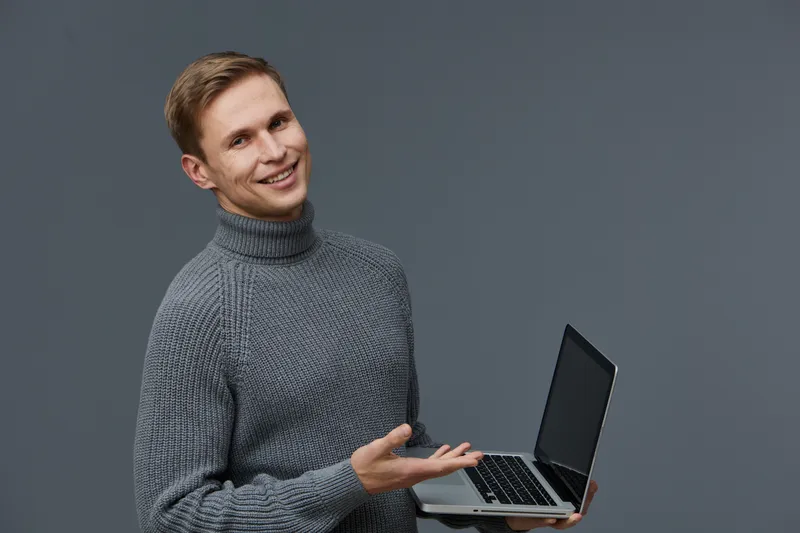 Smiling young man holding a laptop in his hand, wearing a cozy gray turtleneck sweater, looking confidently at the camera. Indoor studio shot with neutral gray background capturing a professional and 