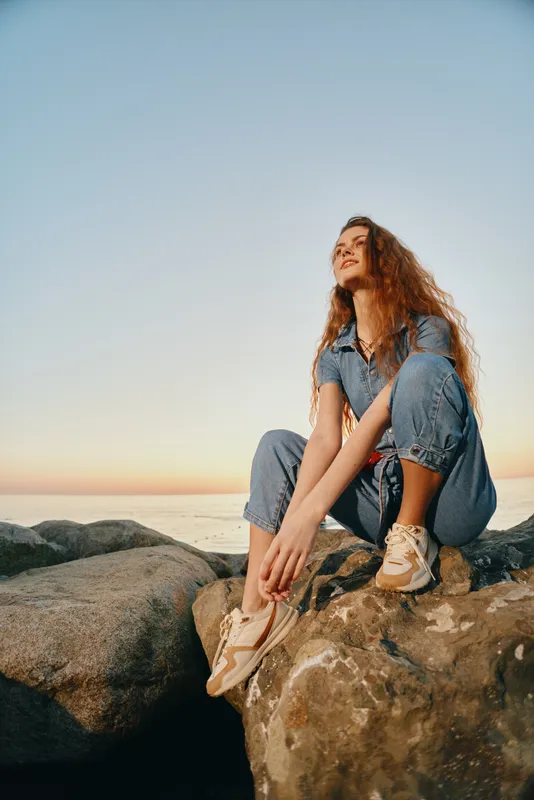 young woman sitting on rocky shore at sunset wearing denim jumpsuit and sneakers, relaxed mood, natural outdoor setting with clear sky