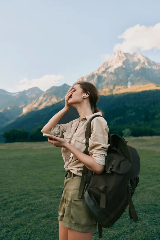 Young woman with backpack standing in a green field with mountains in the background, enjoying nature and outdoor lifestyle during a sunny day.