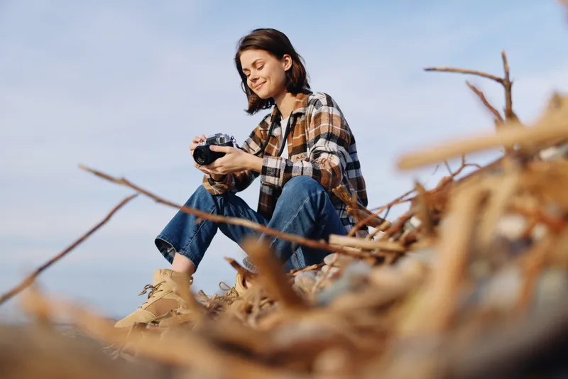 Woman photographer sits among dry stalks in an open field, holding a camera, enjoying a relaxed outdoor moment with casual wear, clear sky, soft light, and natural scenery. Vibrant background adds