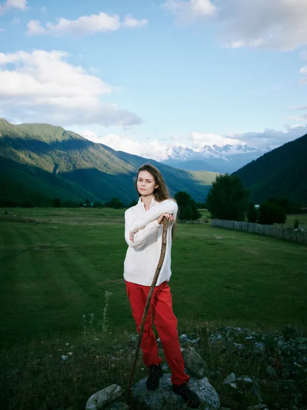 Outdoor scene of a woman standing on rocks in a green valley, holding a walking stick, with distant mountains and blue sky, conveying exploration, serenity and connection with nature