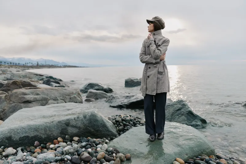 Woman in coat and hat stands on rocky shore looking thoughtfully at calm sea under cloudy sky. Concept of contemplation, solitude, and peaceful nature by the water.