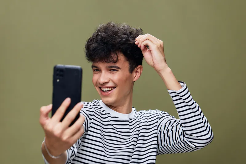 young man taking selfie with smartphone smiling, wearing striped shirt on plain olive background, casual style and joyful expression, modern technology use