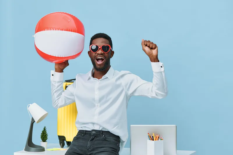 Joyful young man in casual business attire holding a colorful beach ball, celebrating success at the office with a cheerful expression, isolated on a blue background, concept of achievement and happin