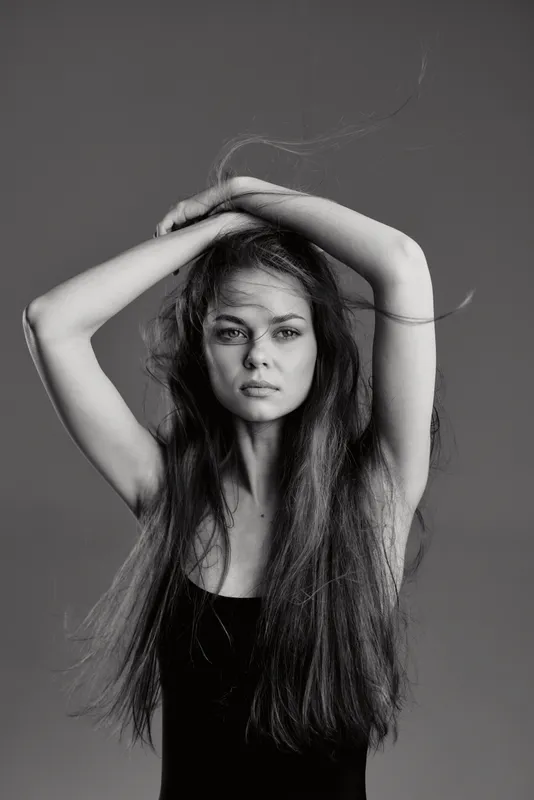 Black and white portrait of a serious woman with long hair posing with hands above head against plain background.