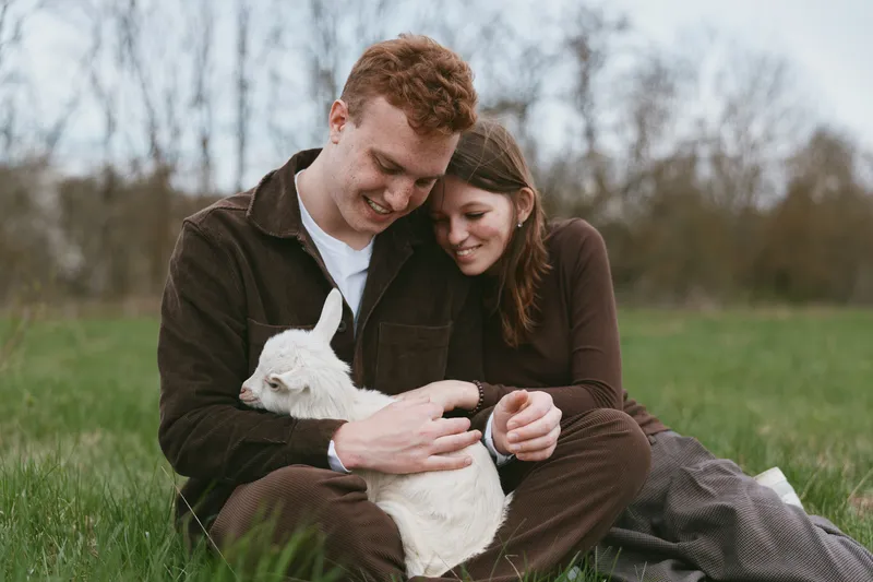 Young couple sitting on grass in casual clothes gently holding a white baby goat outdoors in a natural setting during a relaxed lifestyle moment.