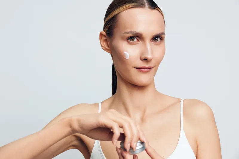 Young woman applies moisturizing cream on her face holds jar with skincare product looks at camera with calm expression isolated on light gray background in studio.