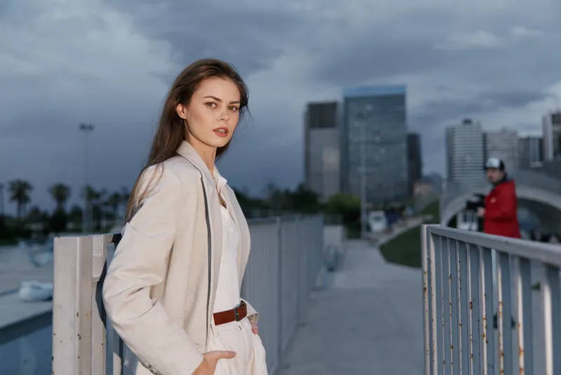 Young woman with serious expression in beige outfit posing outdoors in urban environment under cloudy sky, fashion style, autumn season