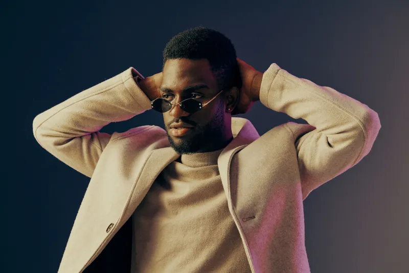 Stylish man in fashionable coat and sunglasses, expressing confidence against a vibrant gradient background, ideal for seasonal fashion concepts