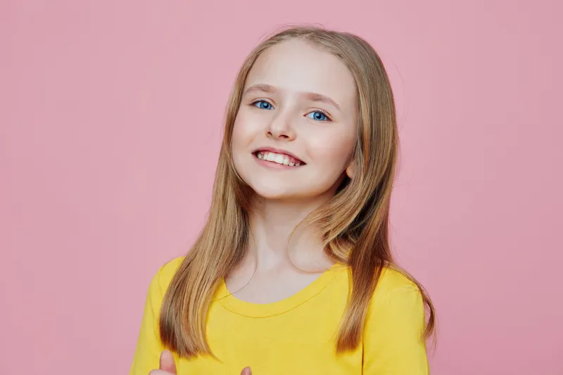 Young girl with long blonde hair smiling in a bright yellow shirt against a pink background, showing joy and positivity