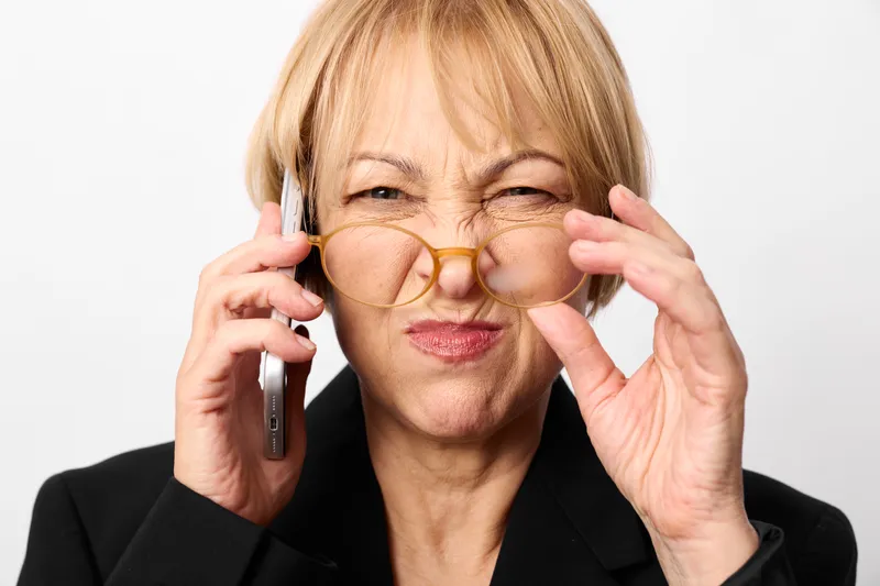 Senior woman wearing glasses adjusting frame, holding phone to ear, smiling, studio white background, concept of vision care and communication, soft light adds confidence
