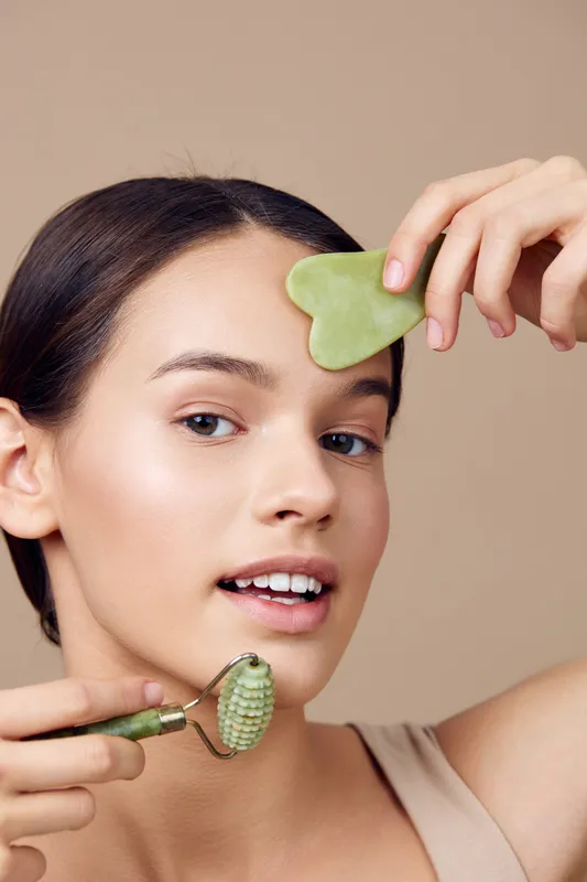 Young woman using a jade roller and gua sha for skincare, showcasing a natural glow and healthy complexion against a soft beige background