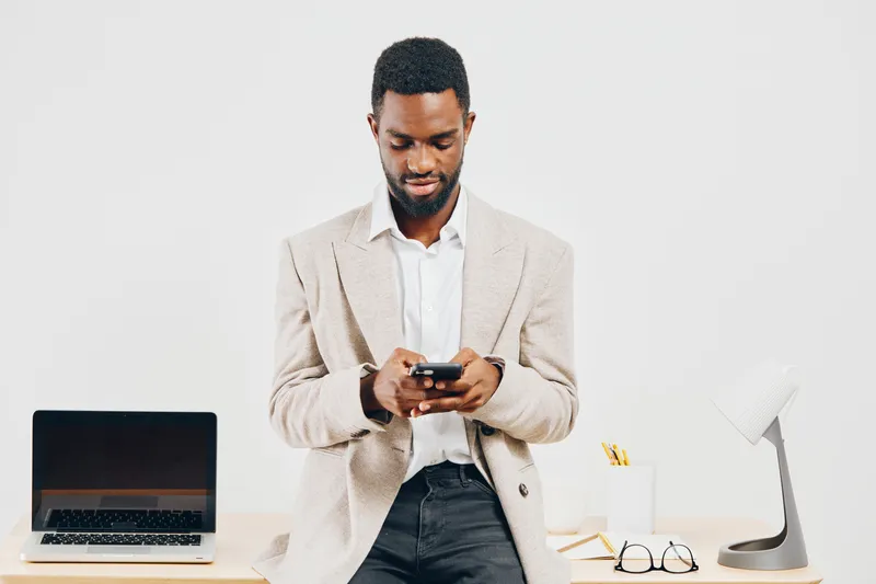 Confident man using smartphone in modern office setting with laptop, desk lamp, and glasses, dressed in smart casual, focused on device, working or communicating, indoor workspace, professional atmosp