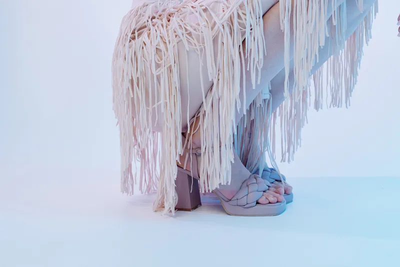 Close-up of a woman wearing stylish sandals and a fringed dress. The image focuses on fashion, beauty, and summer style with soft lighting on a light background.