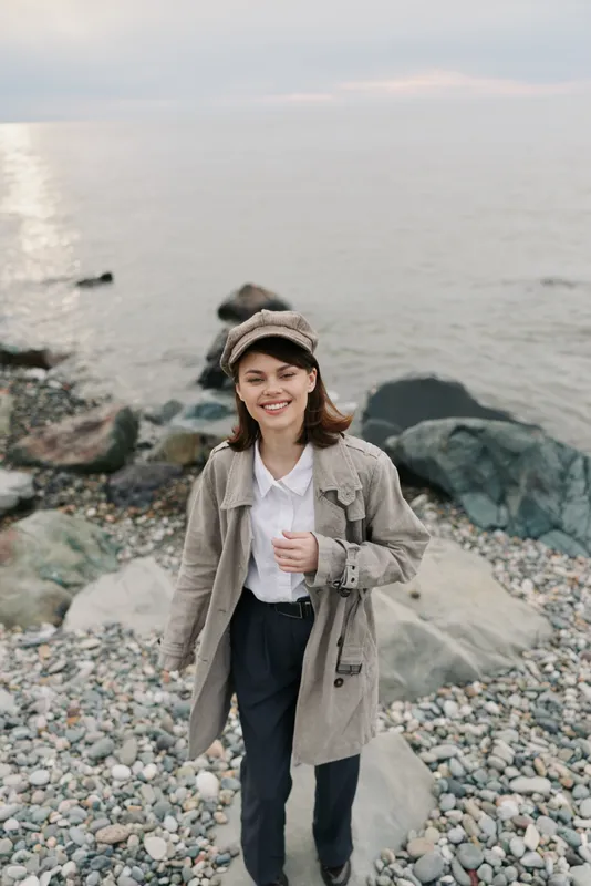 Smiling young woman wearing beige coat and hat stands on rocky beach near calm sea during cloudy day. Concept of casual fashion, nature, outdoor lifestyle, and happiness.