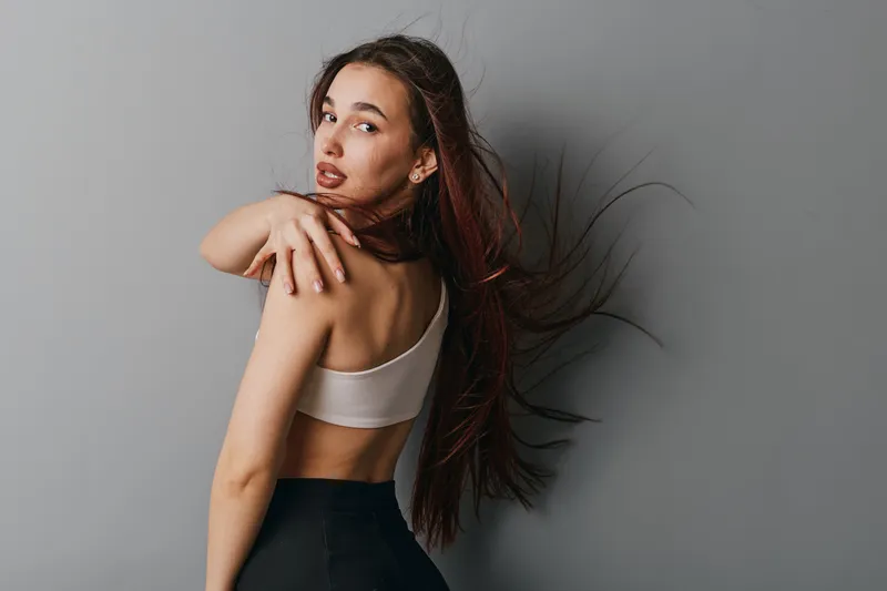 Woman with long hair and bare back posing in sportswear, looking over shoulder on gray background. Fitness, beauty, health and lifestyle concept with natural makeup and casual style.
