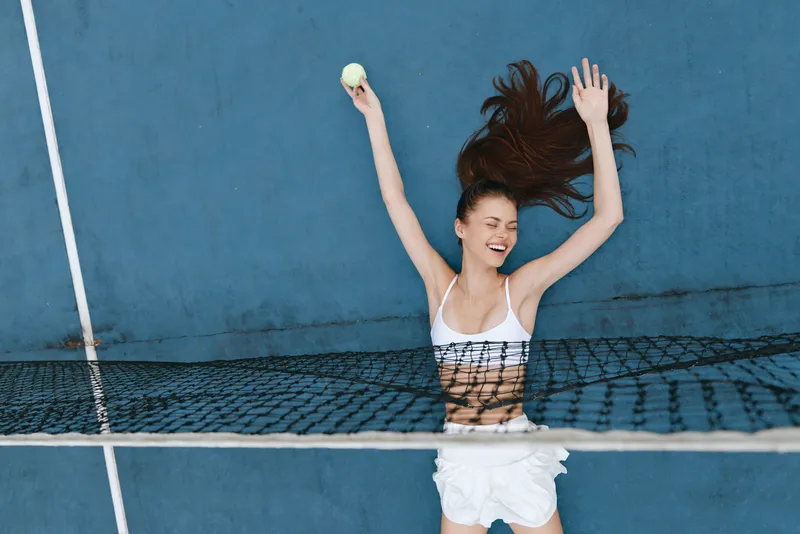 Joyful young woman playing tennis, smiling with her arms raised, wearing sporty attire, against a vibrant blue background, conveying energy and enthusiasm in a summer setting