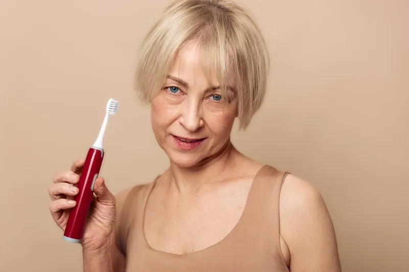 Mature woman with short blonde hair holds red electric toothbrush against beige background. Concept of dental care, oral hygiene, healthy lifestyle, and personal grooming for seniors.