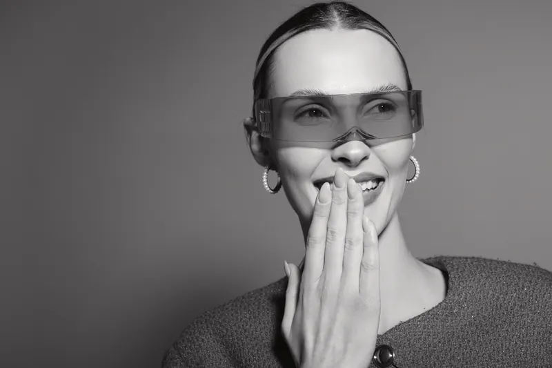 Smiling white woman with light skin wearing transparent glasses and hoop earrings covers mouth with hand in studio. Concept of happiness and casual fashion.