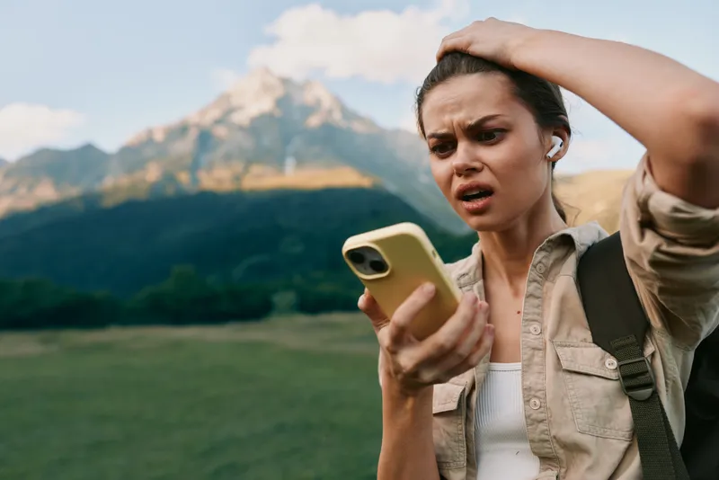Confused woman with earbuds looking at smartphone outdoors in mountains. Concept of technology, communication, and lifestyle. Natural light highlights her concerned expression.