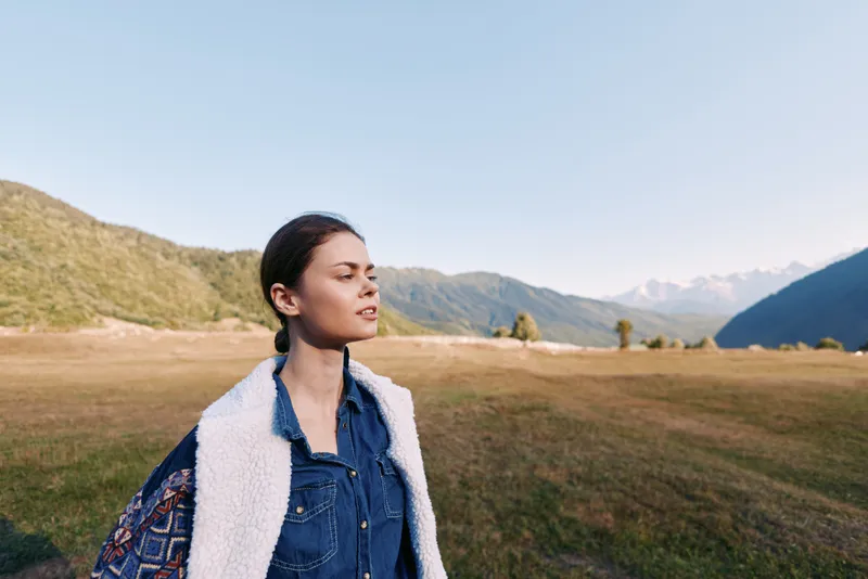 Woman in denim and shearling vest with backpack stands in a sunlit meadow near mountains, portrait with eyes closed enjoying nature, outdoor travel and calm landscape.