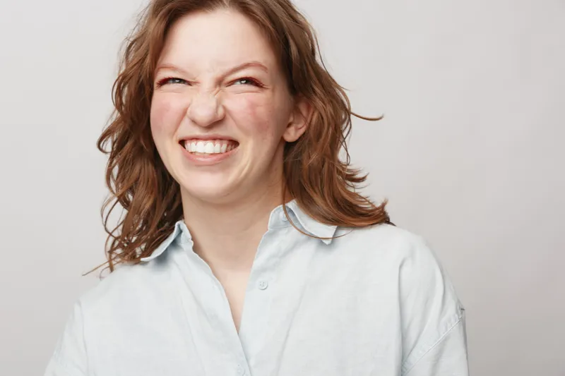 Young woman with red hair smiles broadly showing teeth, wearing white shirt on light background. Concept of happiness, joy, natural beauty, and positive emotions.