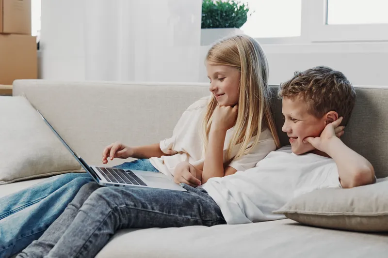 Two children lying on a sofa watch a laptop screen together in a bright living room. The girl and boy share a relaxed moment enjoying digital content and casual home entertainment.