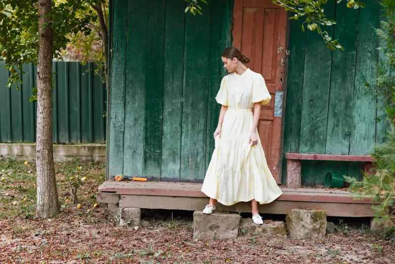 woman dress porch rustic cottage outdoors standing contemplative authentic inclusive portrait of a thoughtful person in a long pale yellow dress on aged wooden steps by a weathered green wall and