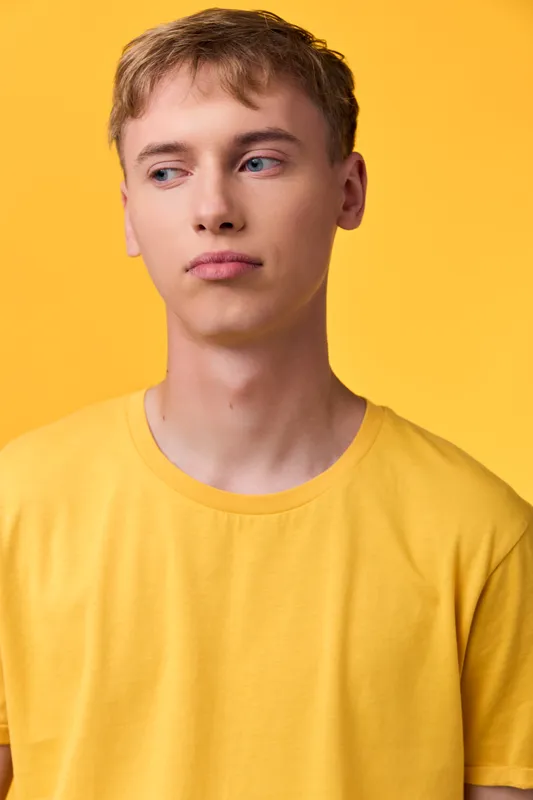Male model wearing a yellow shirt stands against a vibrant yellow backdrop, looking off to the side with a calm, thoughtful expression in a clean studio setting.