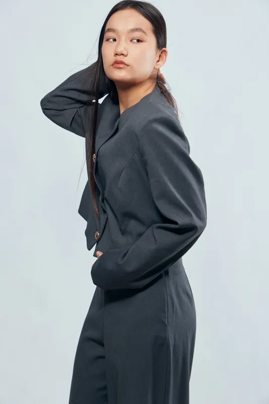 Woman wearing stylish dark gray suit posing with hand touching hair looking aside on light background. Fashion model in modern outfit with confident expression in studio setting.