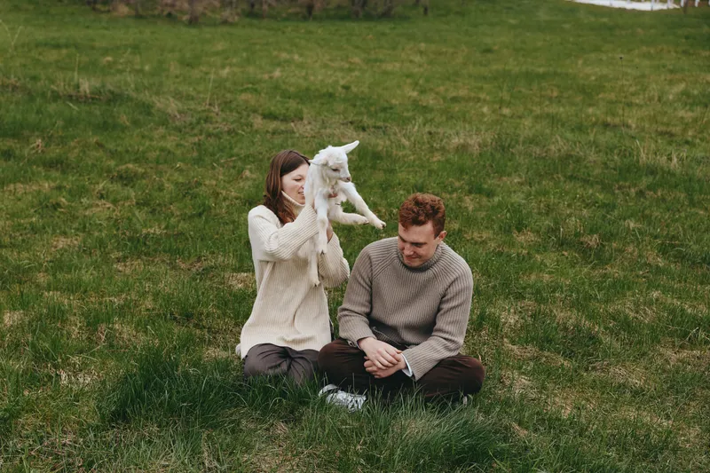A couple sits on grass with a playful kid goat nearby, sharing a tender outdoor moment in a natural meadow, cozy sweaters, relaxed mood, and rustic countryside ambiance.