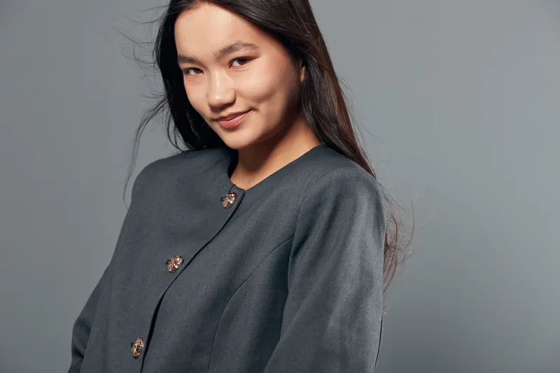 Young woman with long dark hair wearing gray buttoned jacket standing in front of plain gray background smiling confidently with natural makeup and relaxed posture in studio portrait shot.