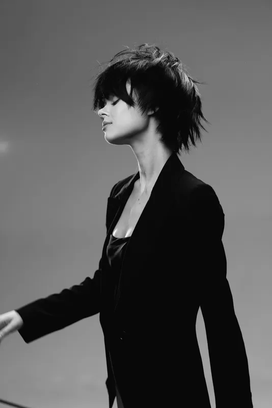 Black and white portrait of a stylish woman with short hair wearing a dark blazer, posing with confidence and artistic flair in a minimalistic studio setting.