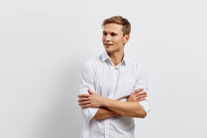 Young confident man with short brown hair wearing white shirt smiling and looking to the side against plain light grey background. Casual style, positive mood, studio portrait. People lifestyle concep